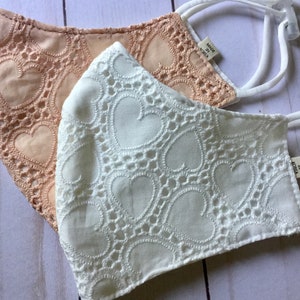 May include: Two face masks with a heart pattern. One mask is peach and the other is white. Both masks have a label that says "Made in Korea".