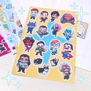 May include: A sheet of colorful stickers featuring cartoon characters in various poses. The stickers are on a yellow background with a blue star design. Other sticker sheets with holographic and colorful designs are visible in the background.