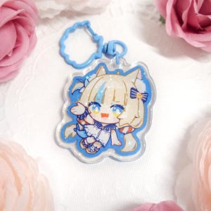 May include: A clear acrylic keychain featuring a cartoon character with blonde hair, blue eyes, and a white and blue outfit. The keychain has a blue border and a blue clasp.