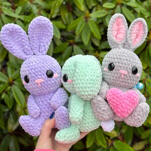 Puede incluir: Tres peluches de conejo de crochet en lavanda, verde menta y gris. El conejo gris sostiene un coraz&oacute;n rosa.