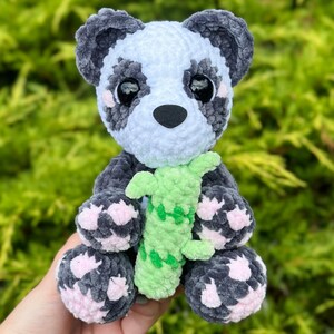 May include: A crocheted panda plush toy holding a green crocheted bamboo shoot. The panda has black and white fur with pink accents on its paws.