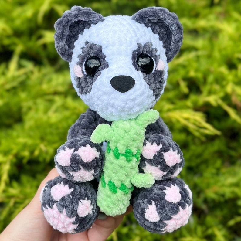 May include: A crocheted panda plush toy holding a green crocheted bamboo shoot. The panda has black and white fur with pink accents on its paws.