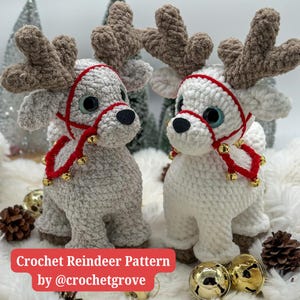 May include: Two crocheted reindeer ornaments, one gray and one white, with red and gold accents. The reindeer are standing on a white fluffy surface with gold bells and pine cones.