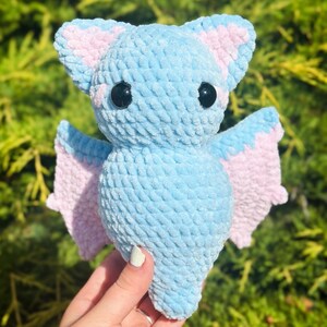 May include: A light blue crocheted bat with pink wings. The bat has black eyes and a fuzzy texture.
