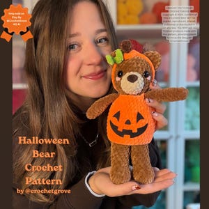 Puede incluir: Un oso de peluche tejido a ganchillo de color marrón que lleva un disfraz de calabaza naranja con una cara de jack-o'-lantern y una pequeña hoja verde en la cabeza. El oso tiene ojos de seguridad negros, nariz marrón y mejillas rosadas.