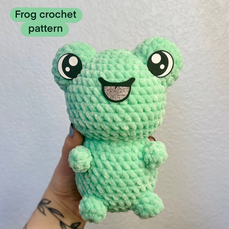 May include: A green crocheted frog with black eyes and a sparkly silver smile. The frog is holding its arms out and has a textured surface.