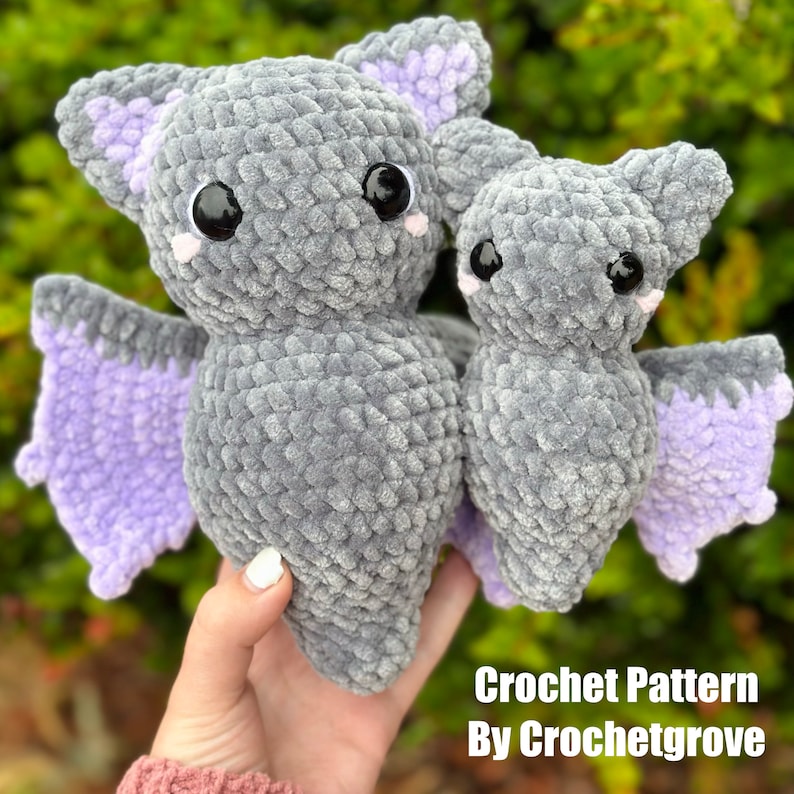 May include: Two crocheted plush bats, one larger than the other, are gray with purple wings and black eyes. The bats are held in a hand. The text "Crochet Pattern By Crochetgrove" is at the bottom of the image.