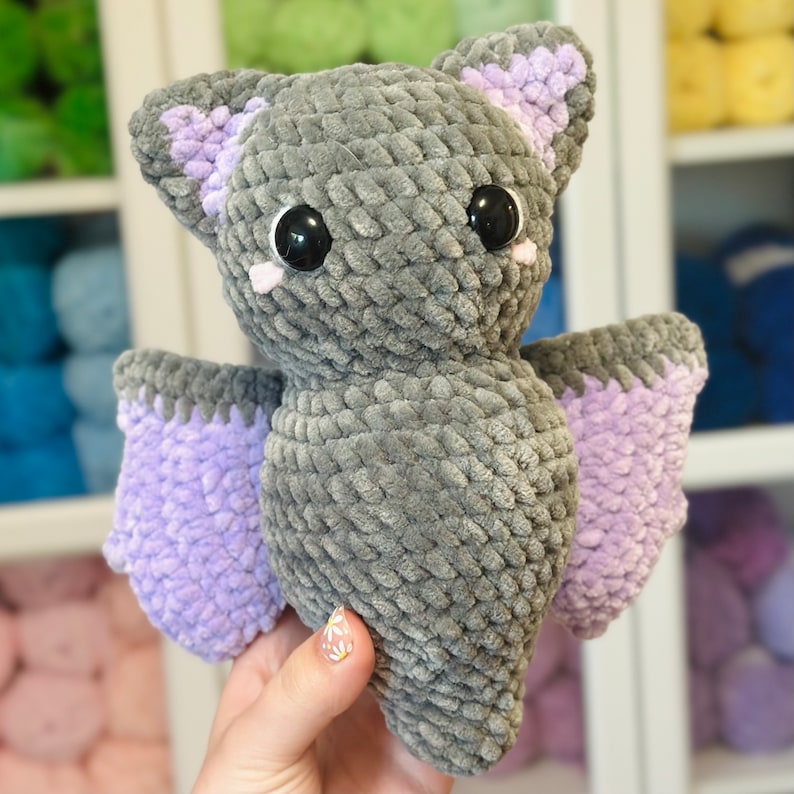 May include: A gray crocheted bat with purple wings. The bat has large black eyes and pink cheeks.