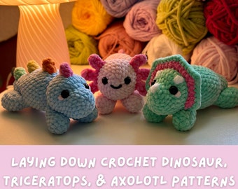 May include: Three crocheted stuffed animals: a blue dinosaur, a pink axolotl, and a green triceratops. The animals are laying down on a white surface. The text "LAYING DOWN CROCHET DINOSAUR, TRICERATOPS, & AXOLOTL PATTERNS BY @CROCHETGROVE" is at the bottom of the image.
