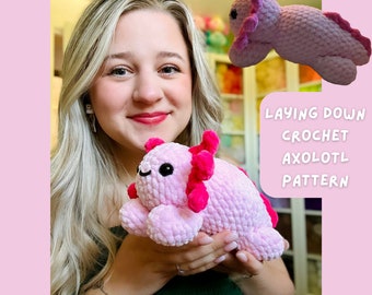 May include: A light pink crochet axolotl plushie with bright pink gills and accents. The plushie is laying down, with the text "LAYING DOWN CROCHET AXOLOTL PATTERN" in white. The creator's handle, "@CROCHETGROVE", is also visible.