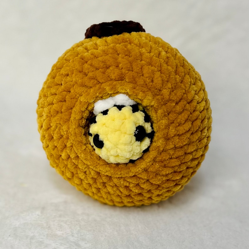 May include: A crocheted bee plush toy with black eyes and a yellow and white body, peeking out of a brown crocheted ball.