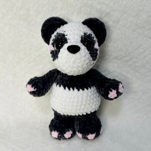 May include: A crocheted panda bear plush toy with black and white fur. The panda has pink accents on its ears and paws.