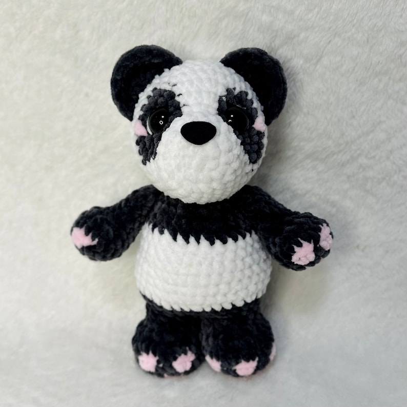 May include: A crocheted panda bear plush toy with black and white fur. The panda has pink accents on its ears and paws.