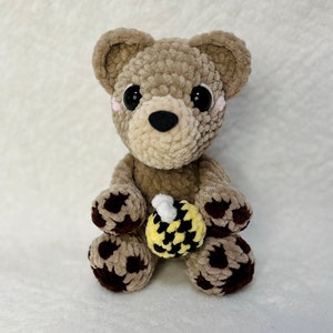 May include: A crocheted plush brown bear with black paw pads holding a yellow and black crocheted bee. The bear has black eyes and a black nose.