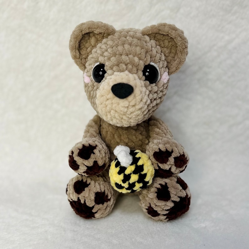 May include: A crocheted plush brown bear with black paw pads holding a yellow and black crocheted bee. The bear has black eyes and a black nose.