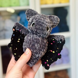 May include: A gray crocheted bat with black wings and colorful accents. The bat has large, black eyes and a fuzzy texture.