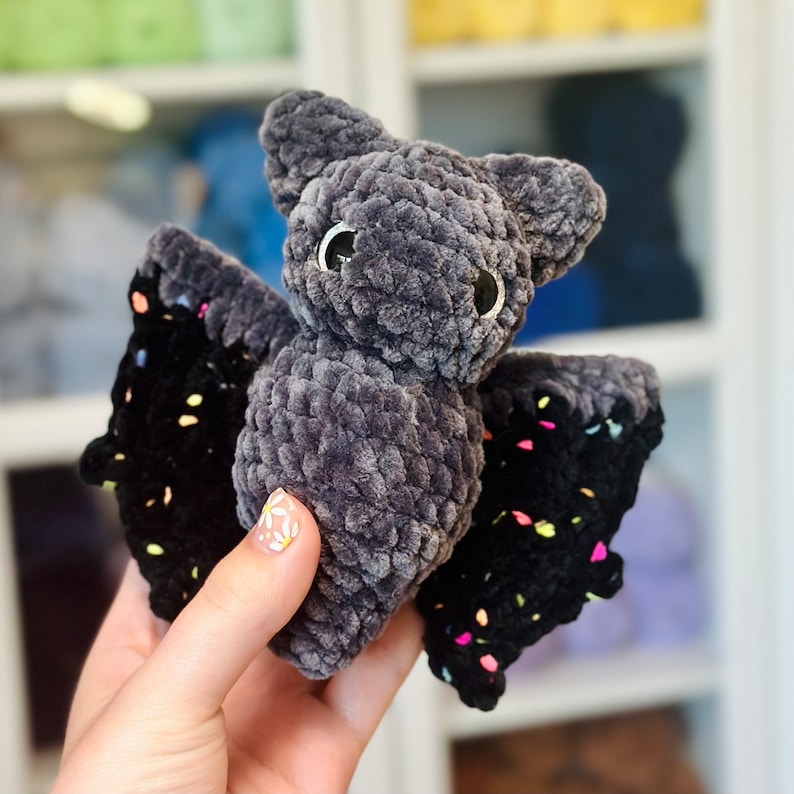 May include: A gray crocheted bat with black wings and colorful accents. The bat has large, black eyes and a fuzzy texture.