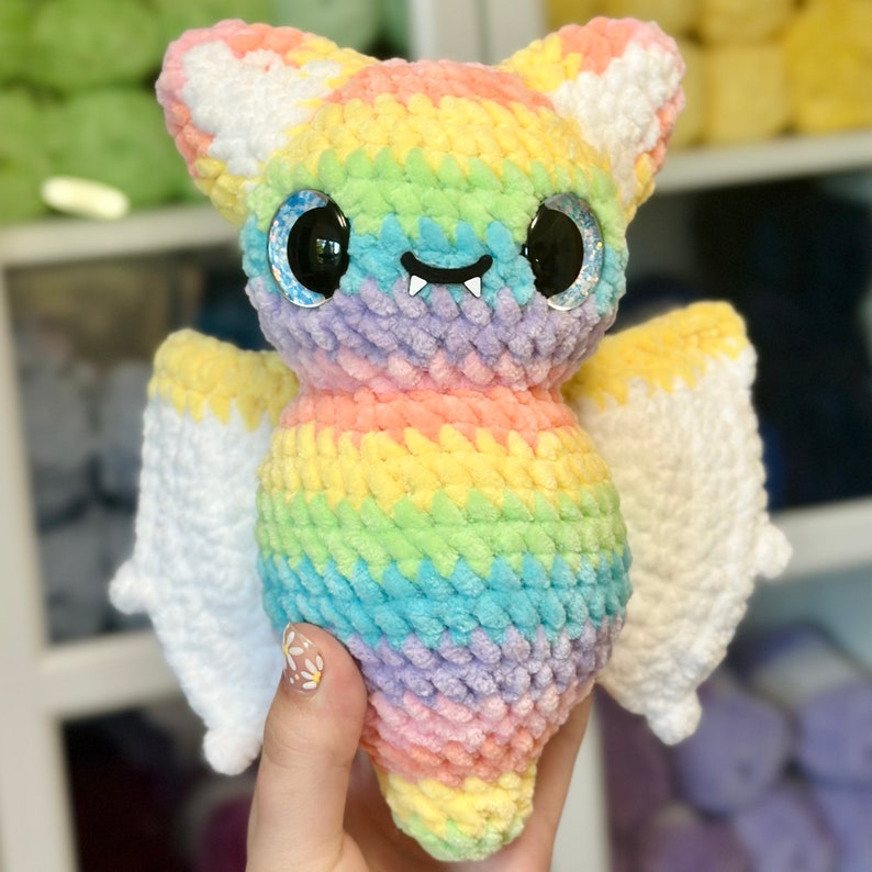 May include: A rainbow crochet bat with sparkly eyes and a toothy grin. The bat has white wings and is made with a soft, fluffy yarn.