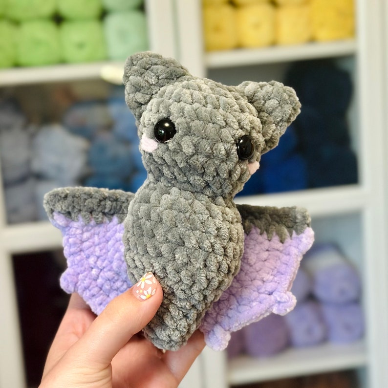 May include: A gray crocheted bat with purple wings. The bat has large black eyes and a pink nose.