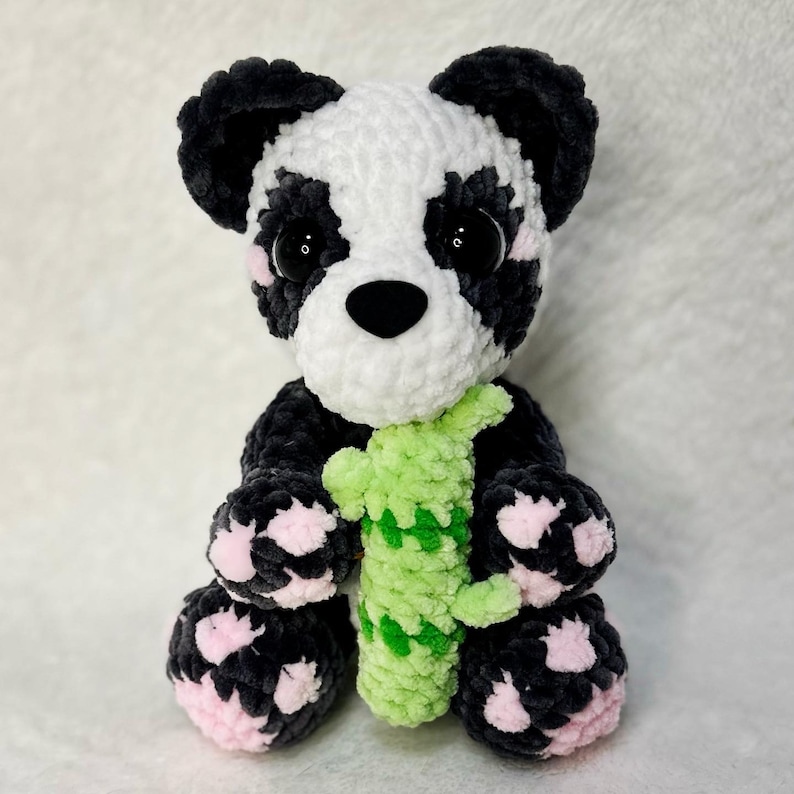 May include: A black and white crocheted panda bear plush toy holding a green crocheted bamboo stalk. The panda has pink accents on its paws and black eyes with pink cheeks.
