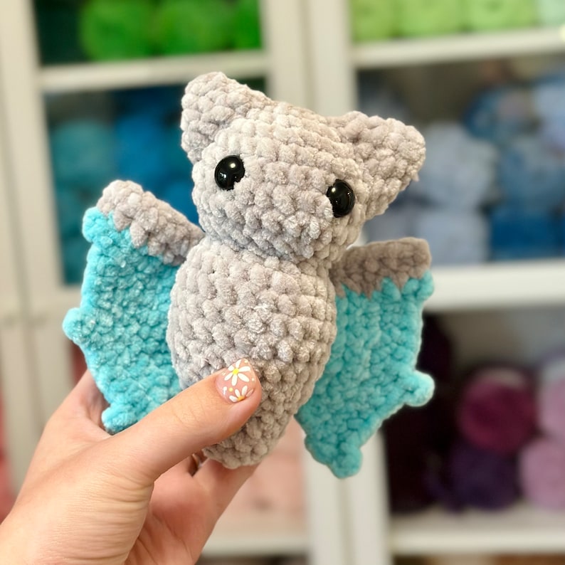 May include: A gray crocheted bat with blue wings. The bat has black eyes and a fuzzy texture.