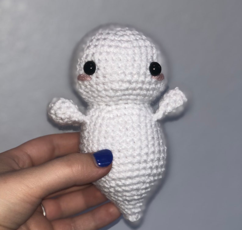 May include: White crocheted ghost plush toy with black eyes and pink cheeks. The ghost has a rounded body and small arms.