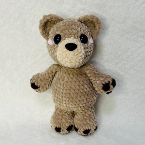 May include: A crocheted brown teddy bear with black eyes and pink cheeks. The bear has brown stitched paws.