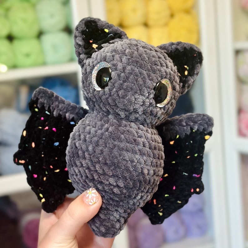 May include: A gray crocheted bat with black wings and colorful accents. The bat has large, sparkly eyes.