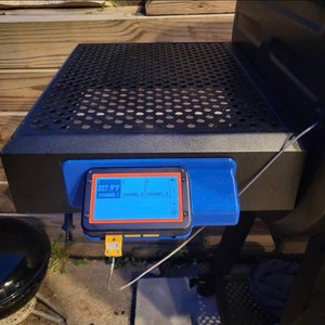 May include: A blue and black digital meat thermometer with a screen displaying "227.9°F" and "CHANNEL 1". The thermometer is attached to a black grill.