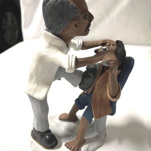 May include: A ceramic figurine depicting a barber shaving a seated customer. The barber, in a white shirt and gray pants, leans over the customer. The customer is seated in a blue chair, wearing a brown jacket and blue pants. The base is white.