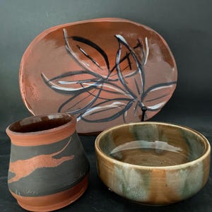 May include: Three handmade ceramic bowls. A large oval bowl with a brown glaze and black and white abstract design. A smaller round bowl with a brown and green glaze. A cylindrical bowl with a brown glaze and a black silhouette of a running dog.