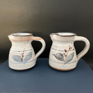 May include: Two small white ceramic jugs with brown and blue leaf designs. The jugs have a rustic, handmade look.