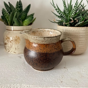 May include: A small, handmade ceramic jug with a rounded body and a handle. The jug features a speckled, light beige rim, a band of orange, and a dark brown base. The jug is displayed on a white surface with two potted plants in the background.
