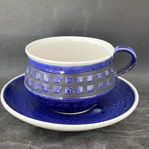 May include: A blue ceramic teacup and saucer set. The cup has a dark blue band with a square pattern, a white interior, and a blue handle. The saucer is solid blue with a white rim.
