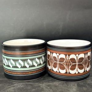 Op de afbeelding: Twee cilindervormige keramische mokken met een witte binnenkant. De linker mok heeft horizontale bruine en mintgroene banden met een wit bladvormig patroon. De rechter mok toont bruine en zwarte horizontale strepen met een wit geometrisch bloemmotief.