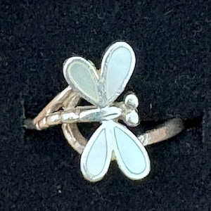 May include: A silver-toned ring featuring a dragonfly motif with iridescent, light-coloured inlay on the wings. The dragonfly body is textured and metallic, with the wings extending outwards from the centre of the band in a symmetrical design.