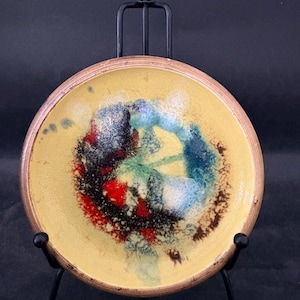 Op de afbeelding: Een ronde keramische schaal met een gele basis en een bruine rand. Het midden heeft een abstract ontwerp met rode, blauwe, groene en zwarte spetters. De schaal wordt gepresenteerd op een zwart metalen ezel.