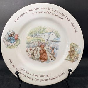 May include: A white ceramic plate featuring a detailed illustration of a girl, a cat, and a farm. The plate has text around the rim: "Once upon a time there was a little girl called Lucie, who lived at a farm called Little-town."