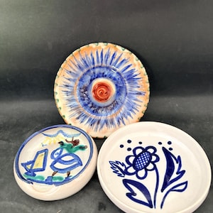 May include: Three ceramic dishes with hand-painted designs. The largest dish has a blue and orange sunburst pattern. The other two are smaller, one with a blue floral design and the other with a whimsical figure.