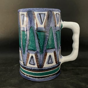 May include: A ceramic mug with a white handle and a cylindrical body. The mug features a blue background with green, brown, and white triangle patterns. A white handle is attached to the side. The base has a green and brown stripe.