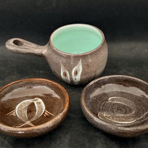 May include: A collection of handmade ceramic bowls and a handled cup. The cup has a light green interior and a brown exterior with a white and brown design. Two small bowls feature bird and swan designs.