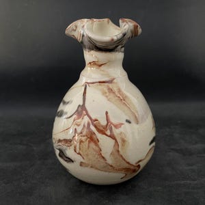 Op de afbeelding: Een kleine, gebroken witte keramische vaas met een bolvormig lichaam en een gerimpelde rand. De vaas heeft bruine en roodbruine abstracte patronen. De vaas heeft een unieke vorm en een getextureerd oppervlak, waardoor het een decoratief stuk is.