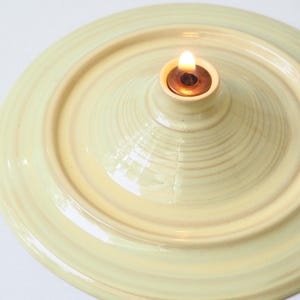 Könnte beinhalten: Eine blassgelbe Keramik-Öllampe mit einem kreisförmigen, abgestuften Sockel und einem zentralen Metallbrenner. Die Lampe hat eine glänzende Oberfläche und eine kleine, brennende Flamme, die aus der Mitte hervortritt und ein warmes Licht abgibt.