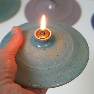 May include: A circular ceramic oil lamp with a light blue crackle glaze and dark speckles. The lamp features a central brass burner insert with a lit flame, creating a warm glow. The surface has a visible network of fine cracks in the glaze.