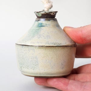 Op de afbeelding: Een kleine, witte keramische olielamp met een bruine rand en een witte lont. De lamp heeft een gespikkelde glazuur en een afgeronde vorm.