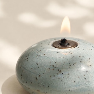 May include: A round, light blue ceramic oil lamp with a speckled finish and a visible crackle glaze texture. The lamp features a central metal burner insert with a small, lit flame extending from the top, creating a warm, glowing light.