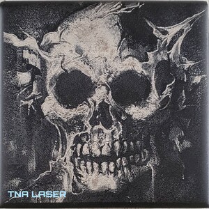 Op de afbeelding: Een zwart-wit afbeelding van een schedel met een getextureerd oppervlak. De schedel is gedeeltelijk bedekt door een donkere achtergrond. De tekst "TNA LASER" is zichtbaar in de linkerbenedenhoek van de afbeelding.