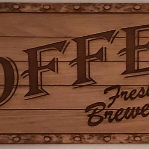May include: A wooden sign with the word "COFFEE" in large, stylized letters. The sign also includes the text "Fresh Brewed 5¢" and a small image of a coffee cup with steam rising from it.