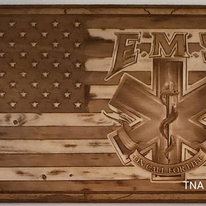 Könnte beinhalten: Eine Holzplakette mit einem verwitterten amerikanischen Flaggen-Design und einem stilisierten medizinischen Symbol mit den Buchstaben "E-M-S" und dem Text "On Call For Life". Die Plakette ist mit "TNA Laser" signiert.