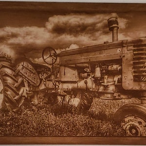May include: A vintage red tractor with a rusted patina sits in a field of tall grass. The tractor has a large front tire and a smaller rear tire. The sky is cloudy and the sun is shining.